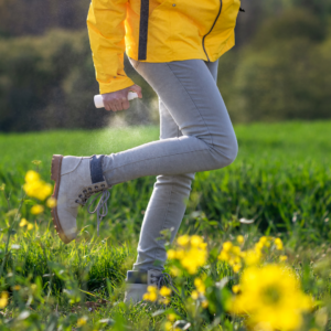 person in yellow jacket, jeans, and hiking boots spraying a substance to prevent tick bites in a field of flowers