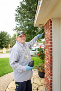 An 855Bugs pest control expert is shown smiling at the camera while treating the exterior of a red brick home.