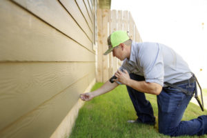 An 855Bugs technician kneels by the side of a beige house inspecting the siding.