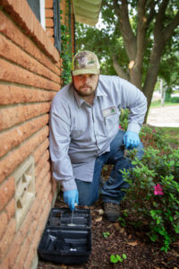 An 855Bugs pest control expert kneels down by the exterior wall of a brick home checking bait traps.
