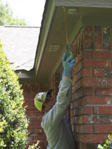 An 855Bugs exterminator is seen stretching up to dust for spiders along the roofline of a brick home.