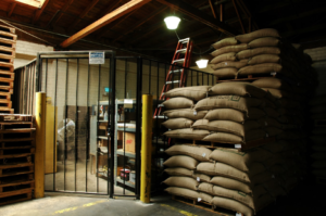 The dimly lit interior of a warehouse is stacked with large bags of grain and supply.